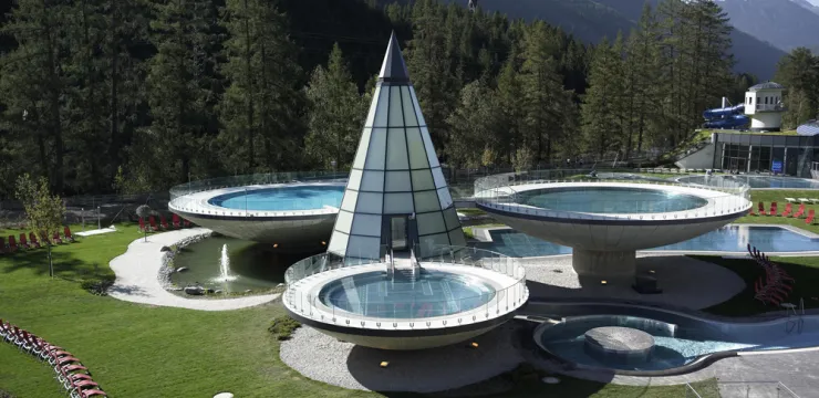 Spitz zulaufendes Glashaus umgeben von drei runden, erhöhten Wasserbecken, Wald und Berge im Hintergrund.