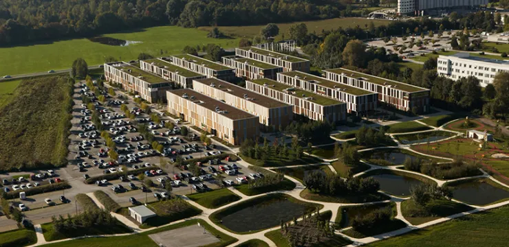 Gebäudekomplex mit begrünten Flachdächern, umgeben von einem Parkplatz und einem Park, Bodensee im Hintergrund.