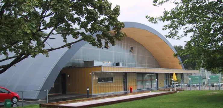 Modernes Gebäude mit gewölbtem Dach aus Metall, umgeben von Bäumen und Rasen.